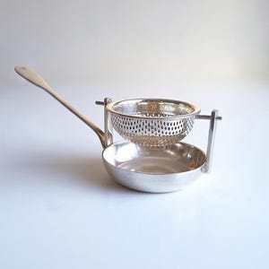 Op de afbeelding: Een zilverkleurige theezeef met een geperforeerde kom die tussen twee verticale steunen aan een ondiepe, massieve basis is bevestigd. De zeef heeft een lange, slanke handgreep aan de zijkant, ontworpen om op een kopje te rusten.