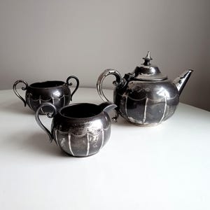 Könnte beinhalten: Ein silbernes Teeservice mit Teekanne, Milchkännchen und Zuckerdose. Das Set hat ein dekoratives Design mit einem gewellten Rand und einem Blumenmuster.