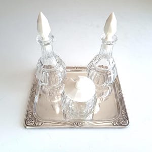 May include: A silver-toned rectangular tray with floral corner detailing holds two clear glass cruets with pointed white stoppers and one small clear glass jar with a matching white lid, all featuring vertical ribbed glass patterns.