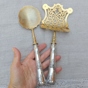 Op de afbeelding: Twee zilveren serveerscheppen met sierlijke handvatten. De scheppen hebben een gouden afwerking en zijn versierd met ingewikkelde bloemmotieven. De scheppen worden in een hand gehouden.