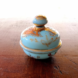 May include: A small, round, blue glass jar with a gold lid and intricate gold and white floral designs. The jar has a second, smaller, round, blue glass lid with gold trim.