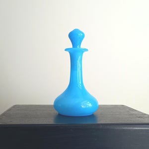 Puede incluir: Una vibrante botella de perfume de vidrio azul claro con un tapón a juego. La botella tiene una base redondeada, un cuello delgado y un tapón decorativo. La botella se exhibe sobre una superficie oscura sobre un fondo blanco.