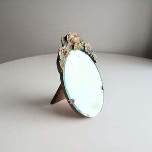 以下が含まれることがあります： 装飾的な花のフレームが付いた楕円形の鏡。 フレームは、ベージュ、緑、青の色合いの彫刻されたバラと葉で飾られています。 鏡は木製のスタンドで支えられており、ヴィンテージ感を演出しています。
