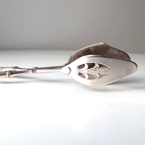 Puede incluir: Un utensilio de servir plateado, posiblemente pinzas, con un diseño floral decorativo en los extremos de la cuchara. El mango tiene una textura similar al bambú. El utensilio está sobre un fondo blanco.