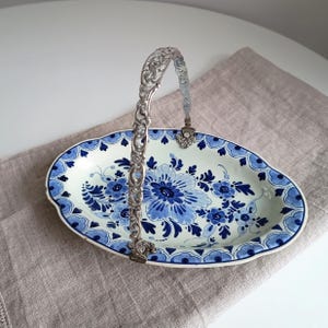 May include: A white oval ceramic serving dish with a blue and white floral pattern and a silver handle. The dish has a scalloped edge and is decorated with a traditional Dutch Delftware design.