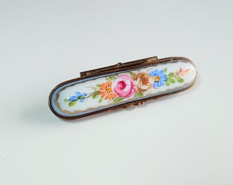 Puede incluir: Pequeño pastillero rectangular antiguo con tapa abisagrada. La superficie de esmalte blanco está decorada con un diseño floral en rosa, naranja, azul y marrón. El marco de metal tiene un aspecto envejecido y deslustrado.