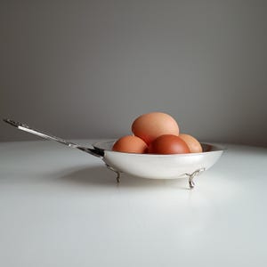 May include: A silver-colored, small frying pan filled with five brown eggs. The pan has a long handle and sits on three small feet. The eggs vary in color from light tan to dark brown.