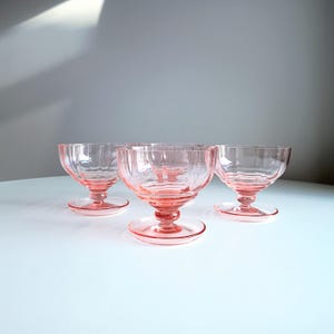 Könnte beinhalten: Vier rosa Dessertschalen aus Glas mit rundem Boden und Stielfuß. Die Schalen haben ein strukturiertes Design. Die Schalen sind auf einer weißen Oberfläche angeordnet.