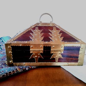Op de afbeelding: Een houten kist met een scharnierend deksel en een metalen handvat. De kist is afgewerkt met messing en heeft decoratieve messing accenten. Het hout heeft een donkerroodbruine kleur. De kist staat op een houten oppervlak.