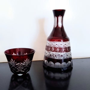 Puede incluir: Un juego de dos decantadores de vidrio rojo rubí con intrincados diseños de vidrio cortado. El decantador más grande tiene una base ancha y un cuello estrecho. El decantador más pequeño es una taza con un borde ancho.