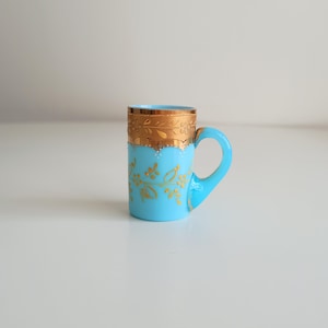Puede incluir: Una pequeña taza azul claro con borde y asa dorados. La taza presenta diseños florales dorados en el cuerpo. La taza es probablemente de vidrio o cerámica y es adecuada para bebidas calientes.