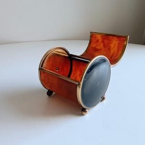 Puede incluir: Una caja de baratijas vintage en forma de barril con tapa con bisagras. La caja presenta un patrón de carey en los lados y la tapa, con una parte delantera de esmalte negro y adornos de metal dorado. Descansa sobre tres pequeños pies esféricos dorados.