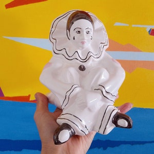 May include: A white ceramic Pierrot figurine, seated with a ruffled collar and black detailing. The clown figure has a teardrop detail and brown hair. The figurine is held against a colourful abstract background.