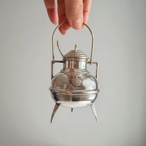 May include: A small, silver metal container with a handle and three pointed legs. The container has a lid and a decorative seashell emblem on the front. The container is held up by a hand against a plain background.