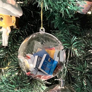 May include: A clear, spherical Christmas ornament filled with miniature books, hanging on a green Christmas tree. The ornament is suspended by a gold string. Other ornaments include a disco ball and a figurine.