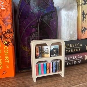 May include: A miniature beige bookshelf filled with tiny books. The books have colorful spines and titles like "Fourth Wing" and "Quicksilver." The bookshelf is on a wooden surface, with larger books and decorative items in the background.