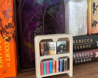 May include: A miniature beige bookshelf filled with tiny books. The books have colorful spines and titles like "Fourth Wing" and "Quicksilver." The bookshelf is on a wooden surface, with larger books and decorative items in the background.