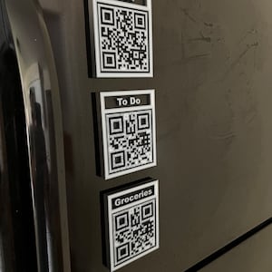 Puede incluir: Tres imanes rectangulares blancos con códigos QR negros y texto en un refrigerador negro. Los imanes dicen "Wi-Fi", "To Do" y "Comestibles".