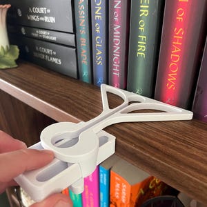 Könnte beinhalten: Ein weißer, 3D-gedruckter Buchhalter in Form eines Notenschlüssels, der zum Einklemmen an einem Regal bestimmt ist. Der Halter befindet sich über einer Reihe farbiger Bücher. Die Bücher tragen Titel wie "Throne of Glass" und "A Court of Mist and Fury".