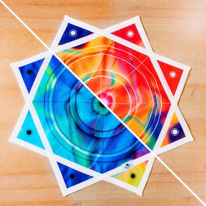 Puede incluir: Una alfombrilla de tela en forma de estrella con un diseño dividido que presenta un degradado azul y verde en un lado y un degradado rojo y naranja en el otro. La superficie muestra patrones circulares concéntricos, bordes cosidos y ojales.