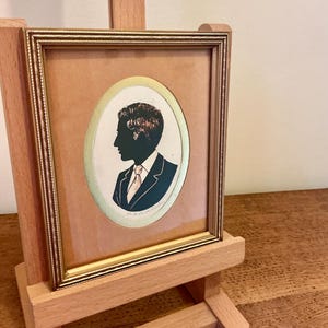 May include: A framed silhouette portrait of a man in a suit and tie, displayed on a small wooden easel. The portrait is in an oval frame with a gold border, set against a tan background. The silhouette is black, with a hint of red in the hair.