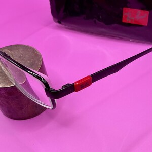 Op de afbeelding: Zwarte en zilverkleurige bril met een rode accent op de poot. De bril heeft een rechthoekig montuur en heldere glazen. Een zwarte etui met een rood logo staat op de achtergrond.