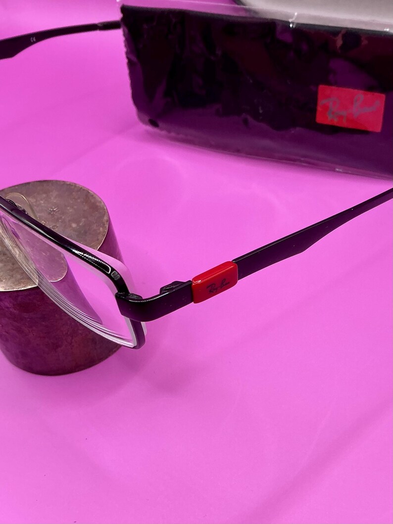 Op de afbeelding: Zwarte en zilverkleurige bril met een rode accent op de poot. De bril heeft een rechthoekig montuur en heldere glazen. Een zwarte etui met een rood logo staat op de achtergrond.