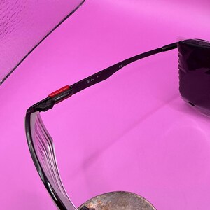 Op de afbeelding: Zwarte zonnebril met een transparant montuur en een rode accent op de poot. Het Ray-Ban logo is zichtbaar op de poot. De bril ligt op een zilveren oppervlak.
