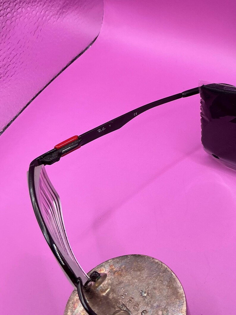 Op de afbeelding: Zwarte zonnebril met een transparant montuur en een rode accent op de poot. Het Ray-Ban logo is zichtbaar op de poot. De bril ligt op een zilveren oppervlak.