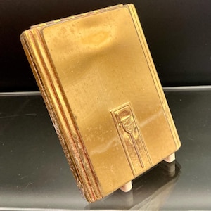 May include: A vintage gold-toned compact with a rectangular shape. The compact features a decorative design on the front and is supported by small, light-colored feet. The edges have a ribbed texture.