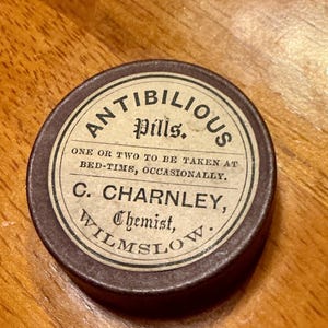 May include: A small, round, antique tin of "ANTIBILIOUS pills." The label is cream-colored with black text, including instructions for use. The tin is brown with a slightly aged appearance, suggesting it is vintage.
