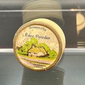 Könnte beinhalten: Eine Vintage-Gesichtspuder-Dose mit rundem Design. Der Deckel zeigt den Text "Grossmith's Face Powder" über einer farbenfrohen Illustration eines Hauses und Bäumen. Auf der Unterseite der Dose steht "Grossmith London".