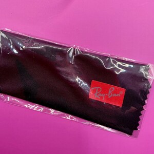 Op de afbeelding: Een zwarte microvezel reinigingsdoek voor brillen, verpakt in een doorzichtige plastic verpakking. De doek heeft een rood Ray-Ban logo. De achtergrond is effen magenta.