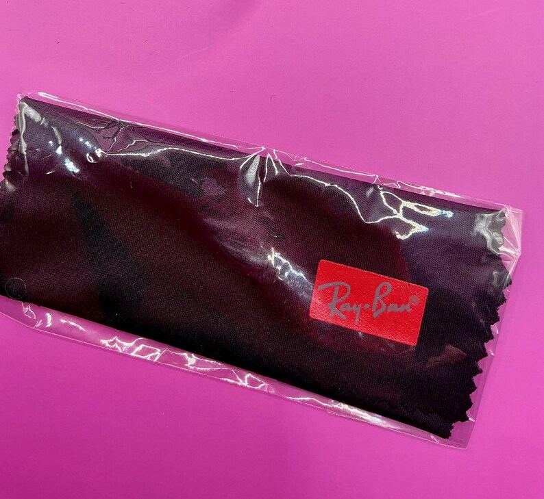 Op de afbeelding: Een zwarte microvezel reinigingsdoek voor brillen, verpakt in een doorzichtige plastic verpakking. De doek heeft een rood Ray-Ban logo. De achtergrond is effen magenta.