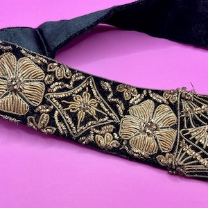 Puede incluir: Cinturón de terciopelo negro con bordado floral dorado. El cinturón presenta intrincados detalles de hilo dorado en un patrón repetitivo de flores y formas geométricas, creando un diseño lujoso y ornamentado. El cinturón está sobre un fondo rosa.