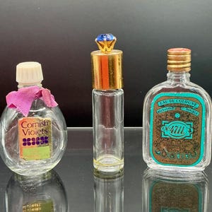 Pode incluir: Três frascos de perfume vintage. O frasco da esquerda tem uma tampa branca, uma fita rosa e um rótulo que diz "Cornish Violets". O frasco do meio é alto e esguio com uma tampa dourada e uma gema azul. O frasco da direita tem uma tampa dourada e um rótulo que diz "Eau de Cologne".