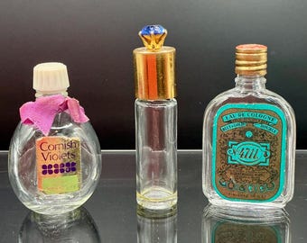 Puede incluir: Tres frascos de perfume vintage. El frasco de la izquierda tiene una tapa blanca, una cinta rosa y una etiqueta que dice "Cornish Violets". El frasco del medio es alto y delgado con una tapa dorada y una gema azul. El frasco de la derecha tiene una tapa dorada y una etiqueta que dice "Eau de Cologne".