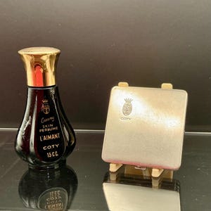 Może przedstawiać: Winietka czarnego flakonu perfum z złotą nakrętką i napisem "Creamy Skin Perfume L'Aimant Coty 15 C.C." obok małego srebrnego kompaktu z logo Coty.