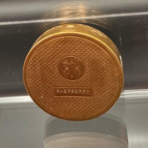 Puede incluir: Un estuche compacto dorado con una superficie texturizada con patrón de diamantes. El estuche compacto presenta la palabra "VALAZE" encima de un logotipo y la palabra "RASPBERRY" debajo. La parte superior del estuche compacto también tiene el nombre "Rubinstein".