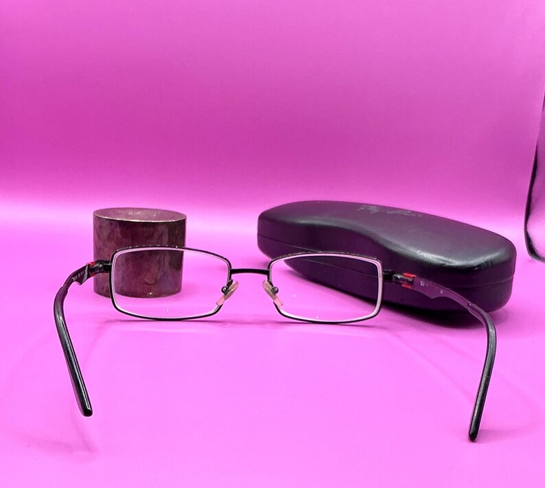 Op de afbeelding: Rechthoekige bril met een zwart montuur en heldere glazen op een roze ondergrond. Een zwarte brillenkoker en een klein, cilindrisch, bruin voorwerp zijn ook aanwezig. De bril heeft een zilveren neusbrug.