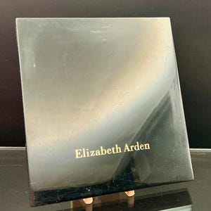 Puede incluir: Un estuche de cosméticos rectangular negro con el texto dorado "Elizabeth Arden" en la parte delantera. El estuche está ligeramente inclinado, revelando una superficie brillante y un reflejo del entorno.