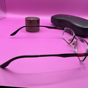 Op de afbeelding: Zwarte en zilverkleurige bril met een rechthoekig montuur en heldere glazen. De bril heeft een rode accent op de veren en staat naast een zwart brillenkoker en een klein, cilindrisch object. De achtergrond is effen roze.