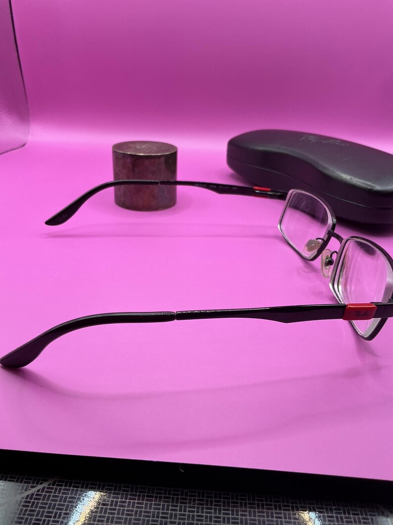 Op de afbeelding: Zwarte en zilverkleurige bril met een rechthoekig montuur en heldere glazen. De bril heeft een rode accent op de veren en staat naast een zwart brillenkoker en een klein, cilindrisch object. De achtergrond is effen roze.