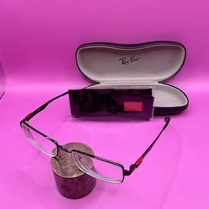 Op de afbeelding: Zwarte en zilverkleurige bril met een rechthoekig montuur en heldere glazen. De bril heeft rode accenten op de veren. Een zwarte etui met het Ray-Ban logo en een zwarte schoonmaakdoek zijn ook aanwezig.