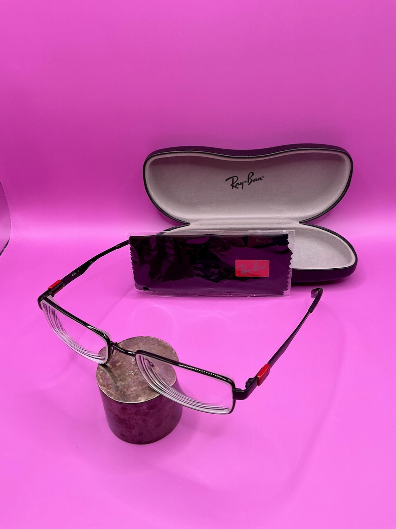 Op de afbeelding: Zwarte en zilverkleurige bril met een rechthoekig montuur en heldere glazen. De bril heeft rode accenten op de veren. Een zwarte etui met het Ray-Ban logo en een zwarte schoonmaakdoek zijn ook aanwezig.