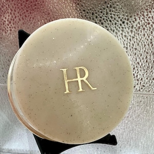 Könnte beinhalten: Ein runder, beigefarbener Kosmetikdöseldeckel mit gesprenkelter Textur und den goldfarbenen Buchstaben HR in der Mitte. Der Deckel ist kreisförmig mit einer glatten Oberfläche und steht auf einem schwarzen Ständer, was ein klares Design zeigt.
