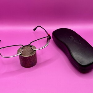 Op de afbeelding: Zwarte rechthoekige bril met heldere glazen en rode accenten op de veren rust op een kleine, cilindrische standaard. Een zwarte brillenkoker met het Ray-Ban-logo staat naast de bril. De achtergrond is effen roze.