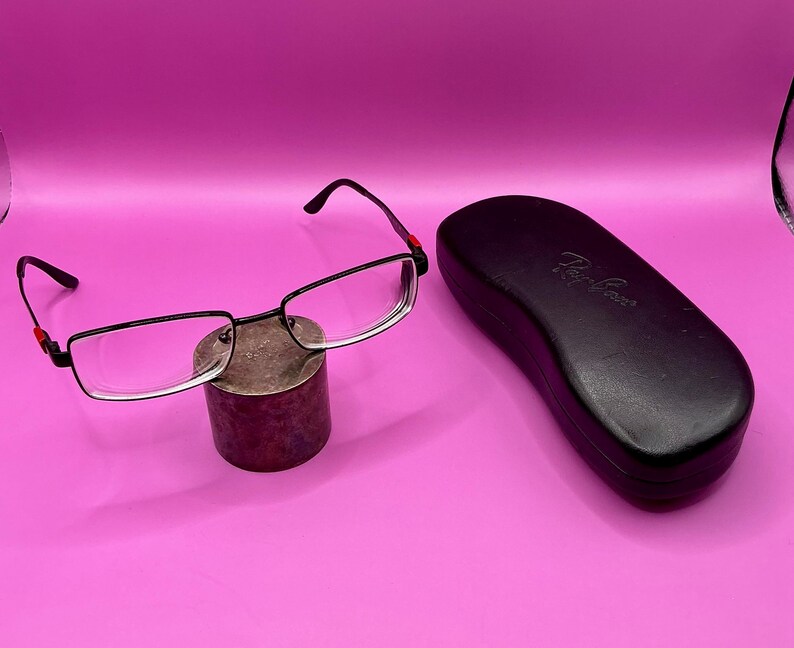 Op de afbeelding: Zwarte rechthoekige bril met heldere glazen en rode accenten op de veren rust op een kleine, cilindrische standaard. Een zwarte brillenkoker met het Ray-Ban-logo staat naast de bril. De achtergrond is effen roze.