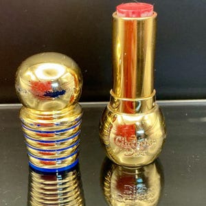 Könnte beinhalten: Eine goldfarbene Lippenstift-Tube und -Kappe. Der Lippenstift hat eine leuchtend rote Farbe. Die Kappe hat eine Kugelform mit blauen und goldenen Streifen. Auf der Lippenstift-Tube steht "Christian Dior".