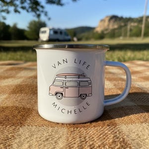 A white enamel mug with a pink campervan illustration and the name Michelle sits on a checkered outdoor blanket.